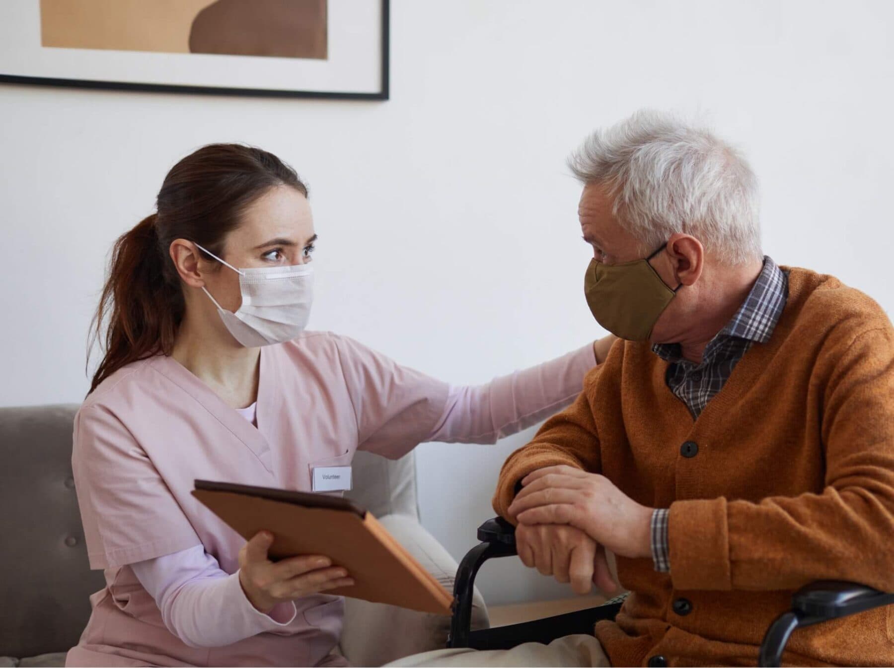Pflegekraft mit Mundschutz spricht mit einem Senior im Rollstuhl und hält eine Mappe in der Hand.