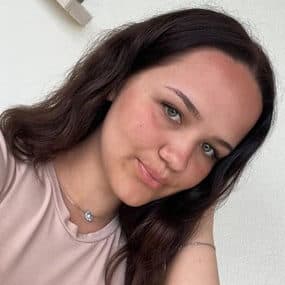 Nahaufnahme-Porträt einer Frau mit langen dunklen Haaren, leichtem Lächeln und hellem Hintergrund im Innenraum.